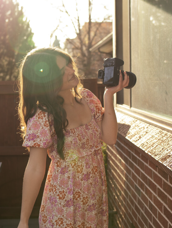A woman with long brown hair wearing a pink floral dress, holding a camera, standing outside on a sunny day next to a brick wall and glass window.