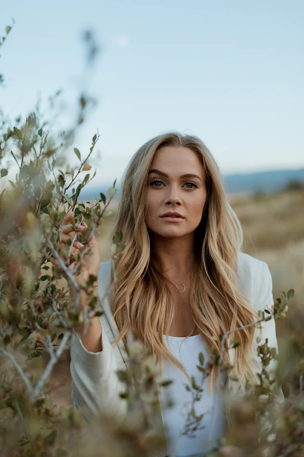 A woman with long blonde hair, wearing a white top and jacket, standing outdoors among green bushes, with a blurred landscape and sky in the background.