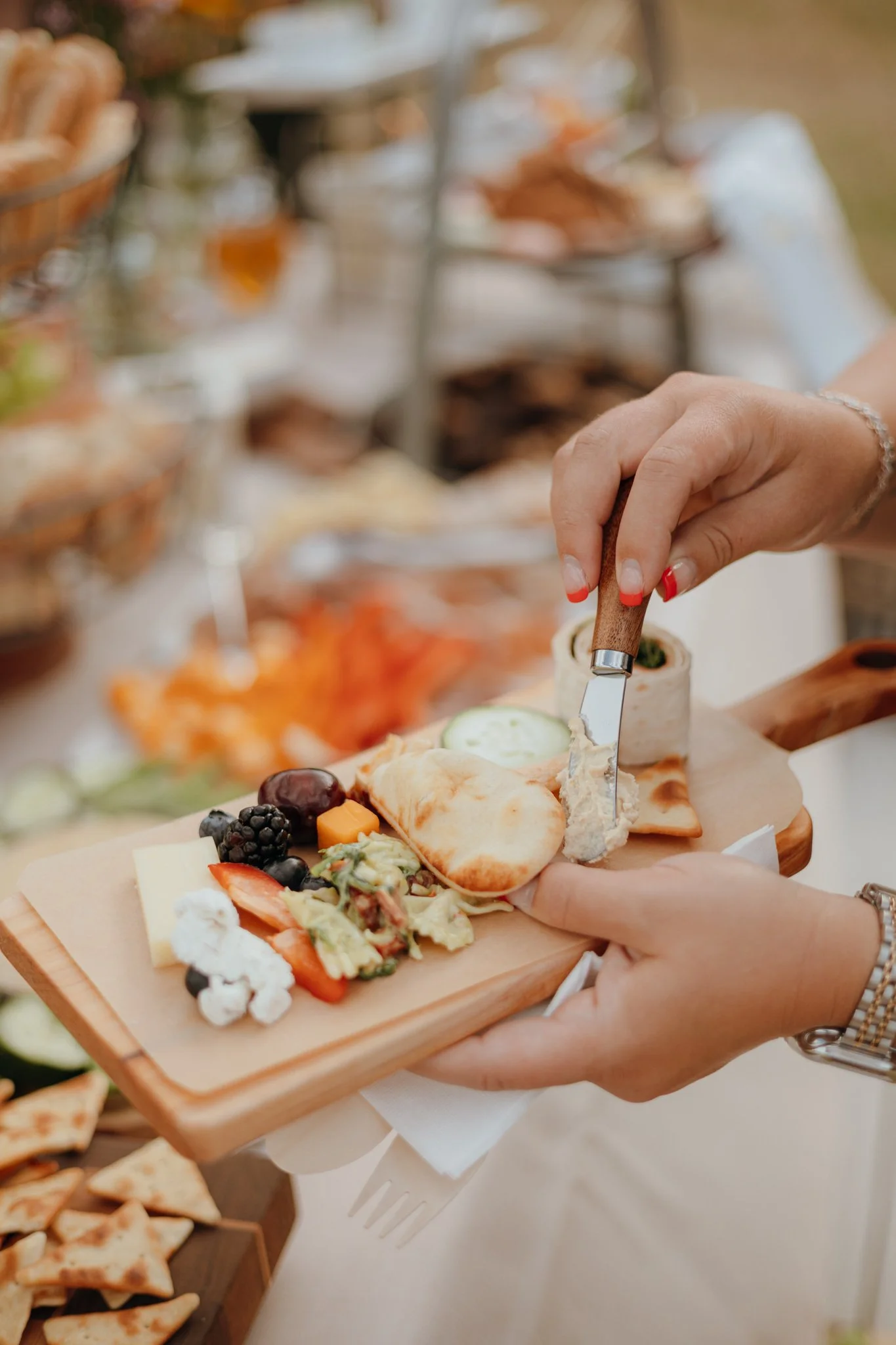 Person serving cheese and crackers with assorted fruits and spreads on a wooden platter at a buffet.