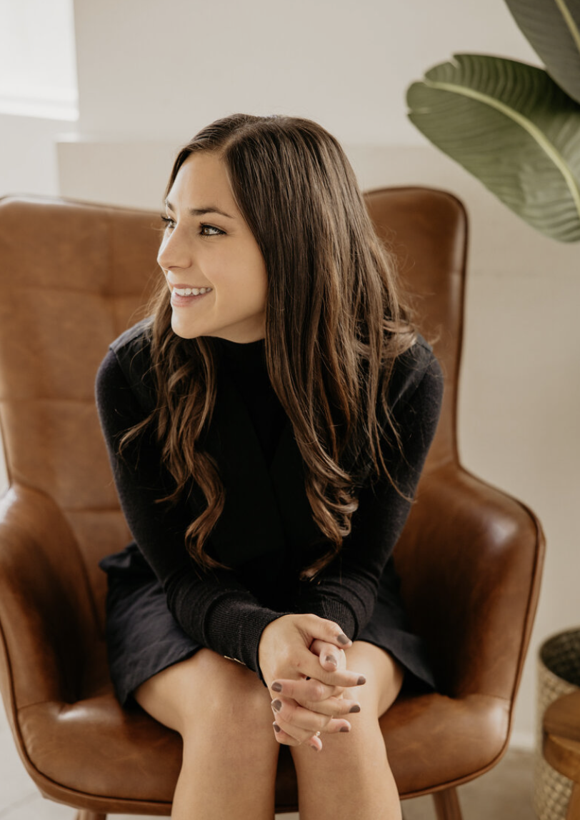 A young woman with long brown hair sitting in a brown leather armchair, smiling and looking to her left, in a well-lit room with a plant in the background.