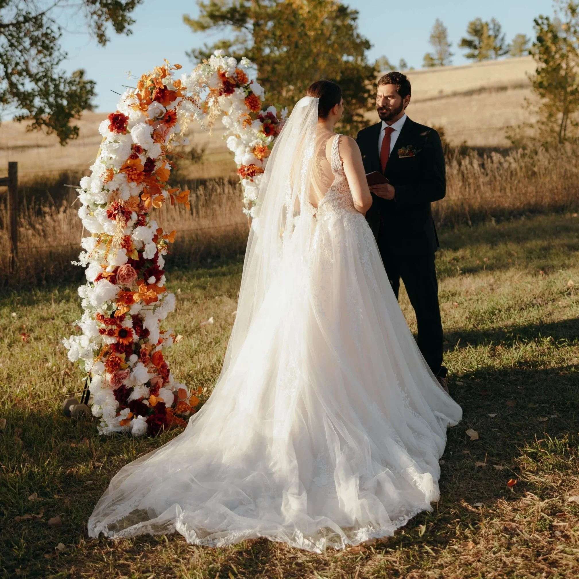 A bride and groom standing outdoors during their wedding ceremony, exchanging vows under a floral arch made of white and orange flowers.