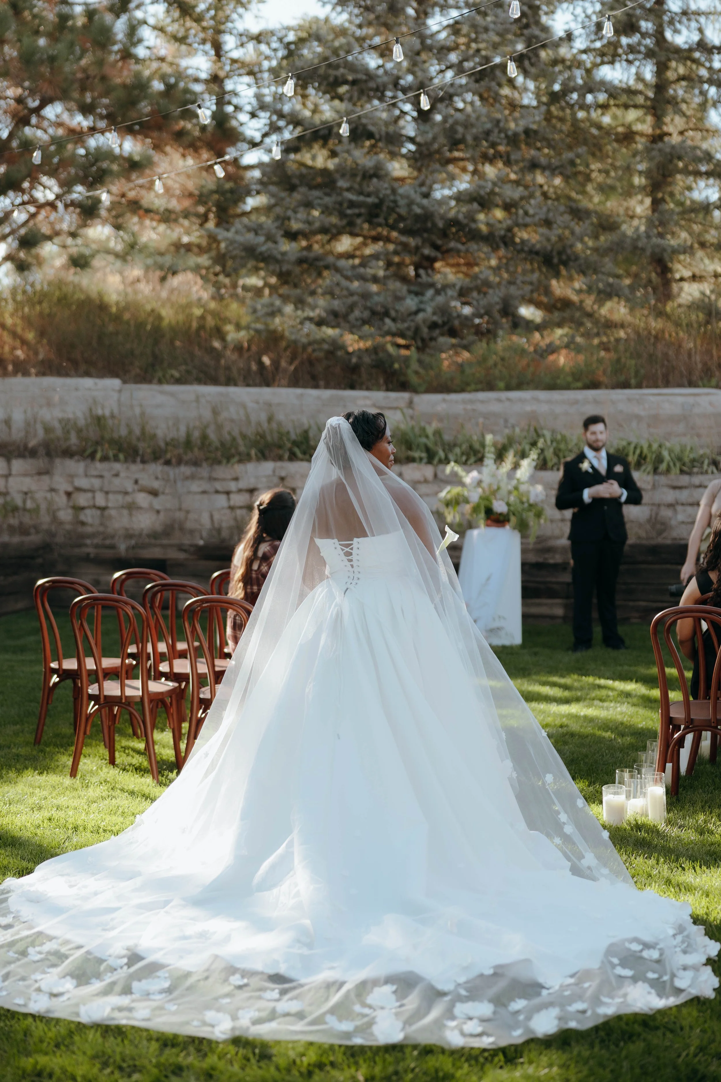 Bride in a white wedding gown and veil standing outdoors during a wedding ceremony, with groom and guests in the background, on a grassy area under string lights, with trees and a stone wall behind.