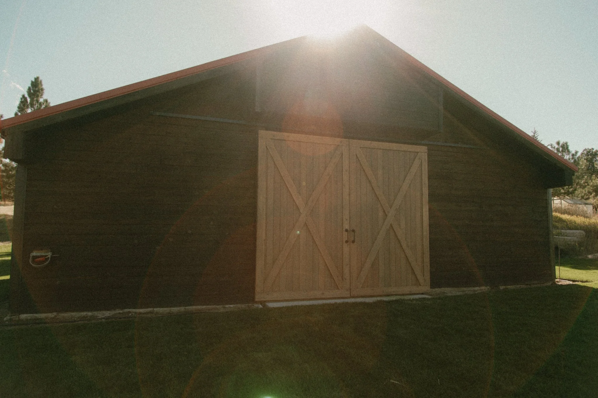 A wooden shed with a red roof and double doors, positioned in a grassy area with sunlight shining from above.