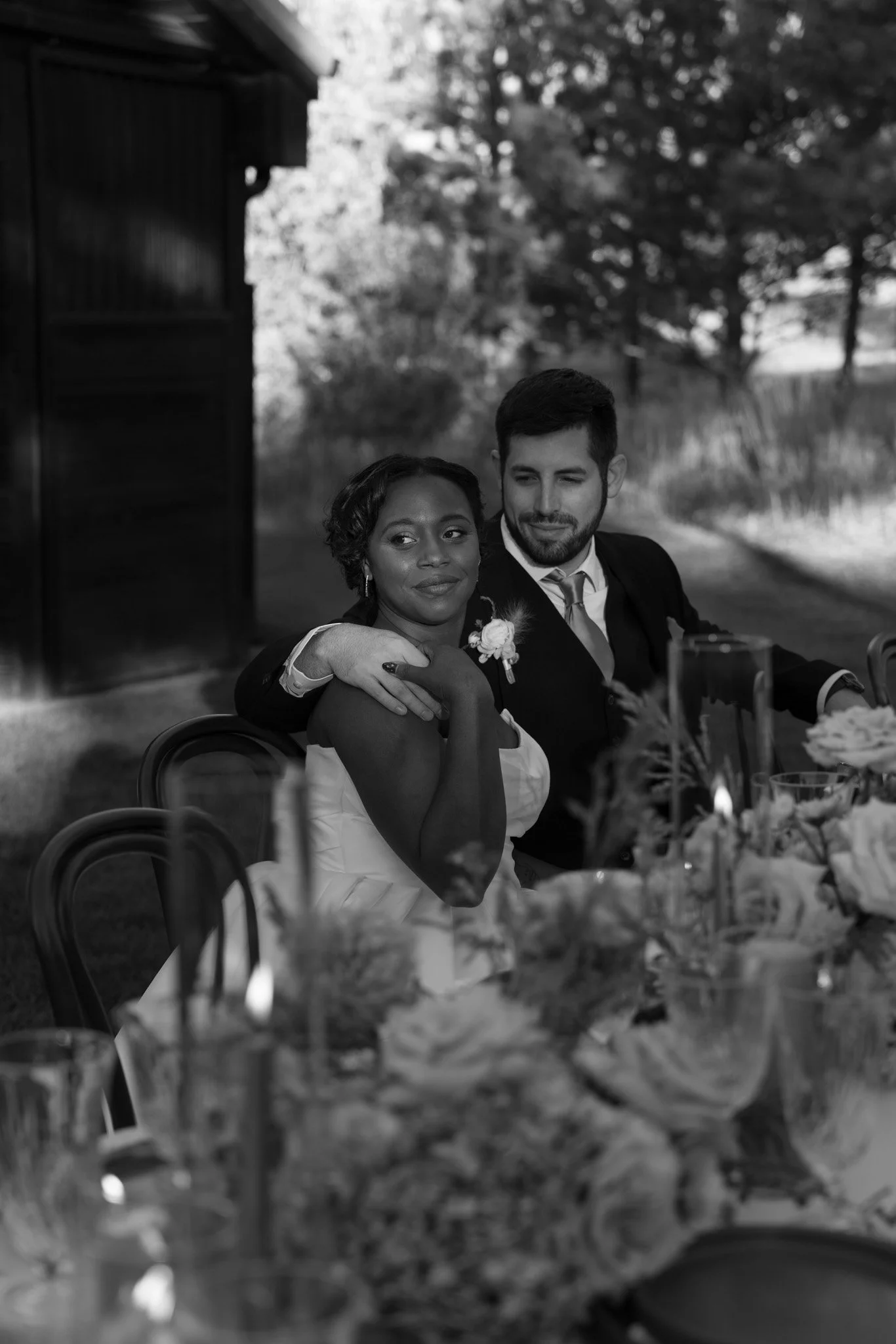 A black and white photo of a couple sitting closely at a wedding reception, with the woman wearing a wedding dress and the man in a suit. They are outdoors, with trees and a barn-like structure in the background. The couple looks content, with the man's arm around the woman.
