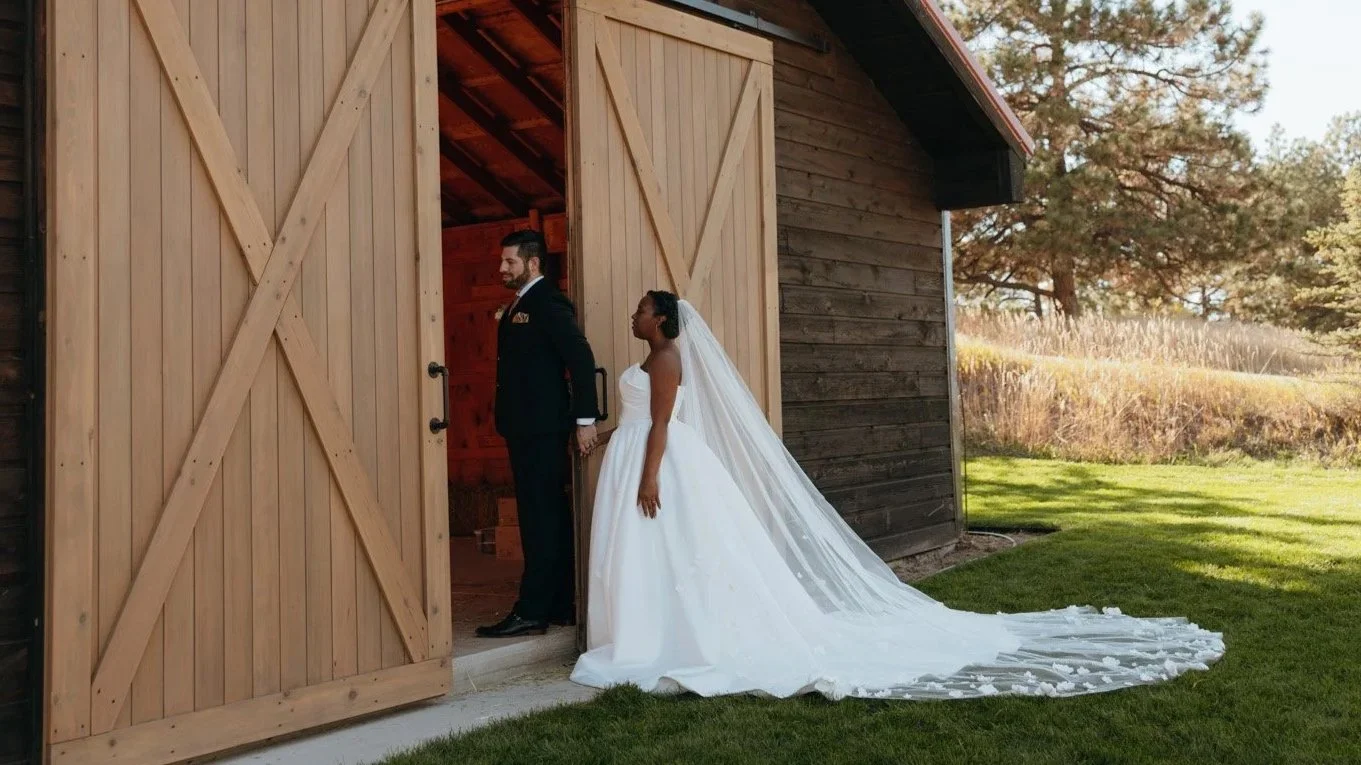 Bride and groom holding hands outside a wooden barn, with the bride wearing a white wedding gown and veil, and the groom in a formal dark suit, during a wedding photoshoot.