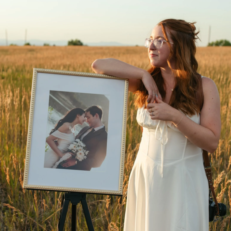 A woman with red hair and glasses stands in a field at sunset, resting her arm on an easel displaying a framed wedding photo of a couple smiling and holding a bouquet.