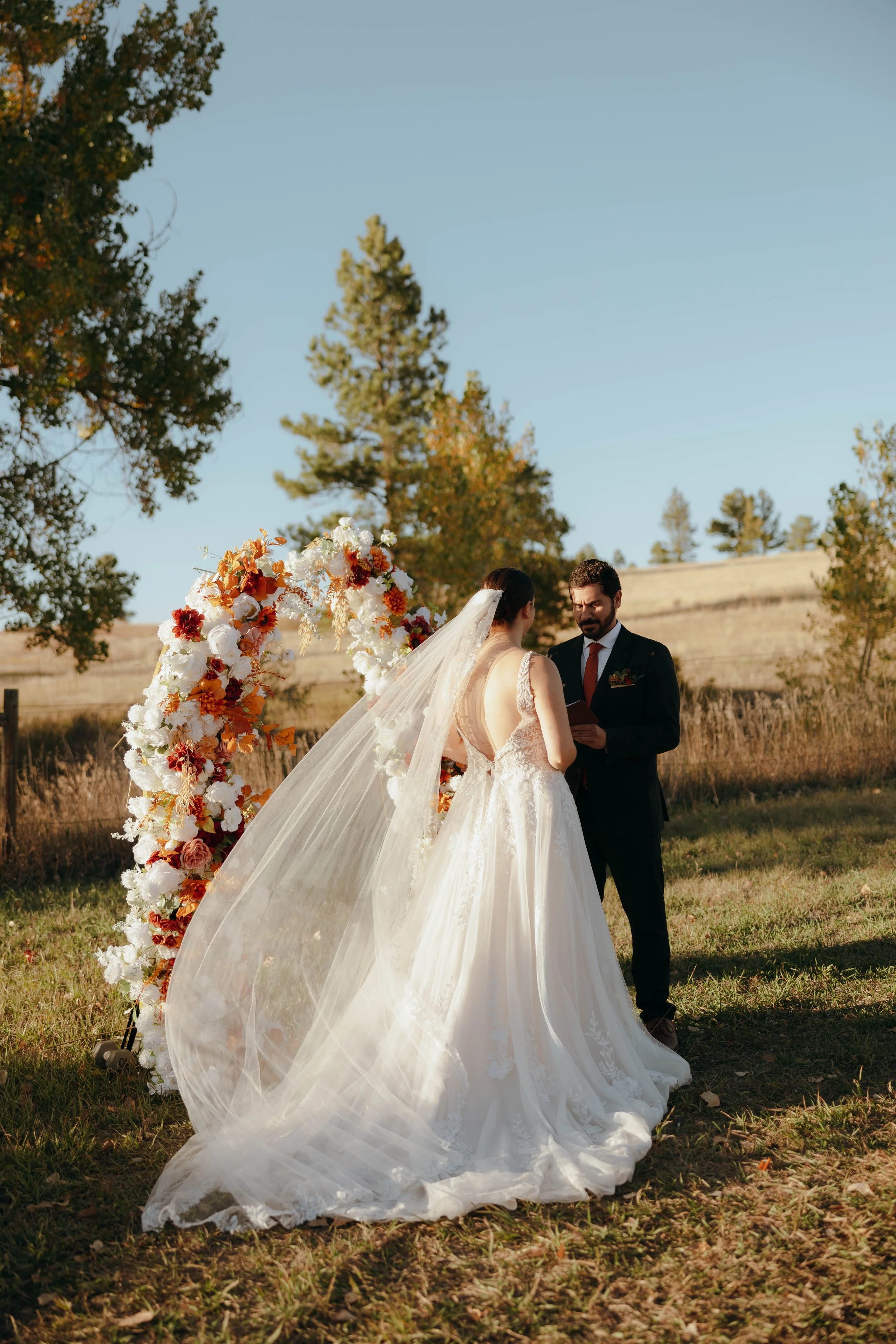 A bride and groom exchange vows outdoors during a wedding ceremony, standing in front of a floral arch with trees and open field in the background.