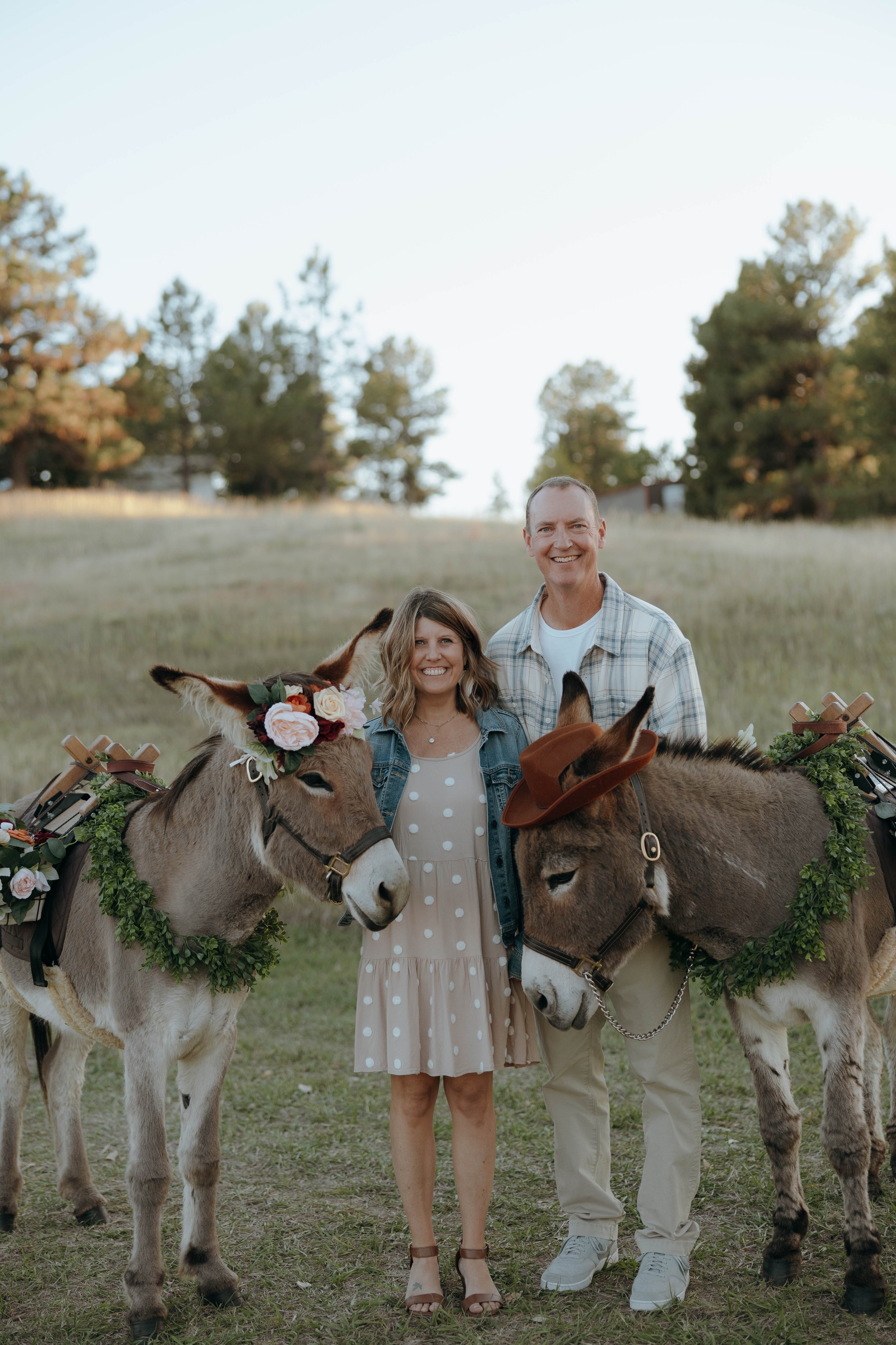 A woman and a man standing outdoors in a field with two decorated donkeys, smiling at the camera.