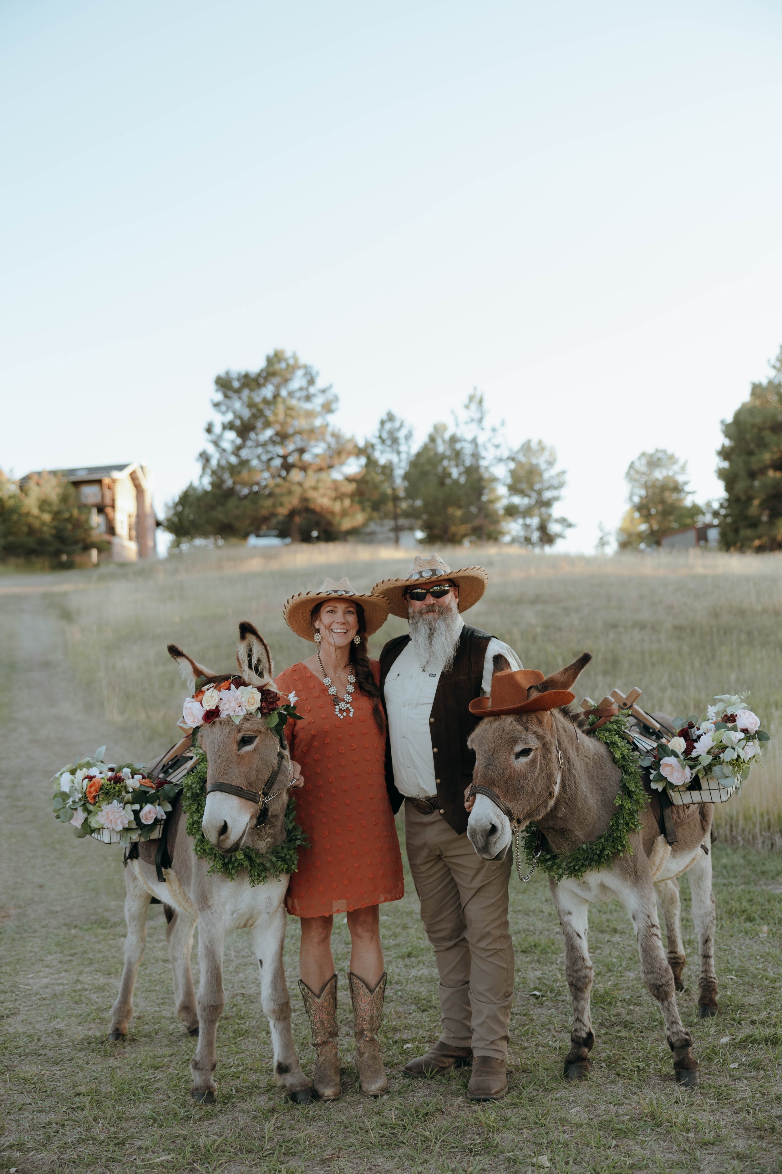A smiling woman and man standing with decorated donkeys in a grassy field with trees in the background, wearing cowboy hats and outdoor attire.