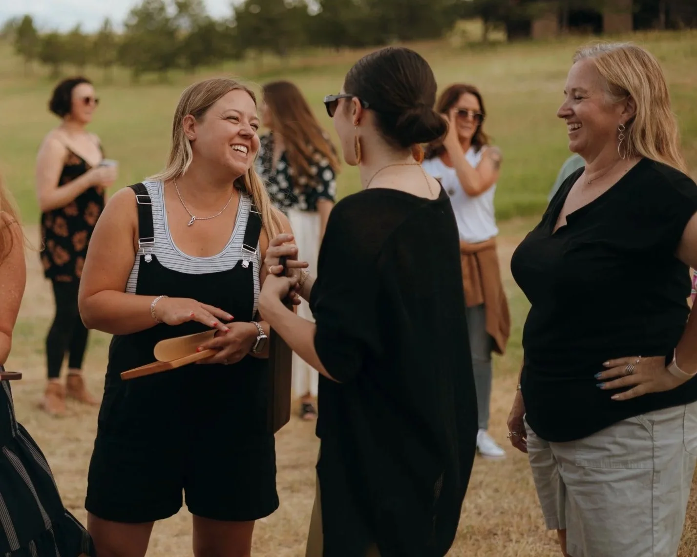 Group of women outdoors, talking and smiling, with some holding drinks and a wooden tray, in a grassy area with trees in the background.