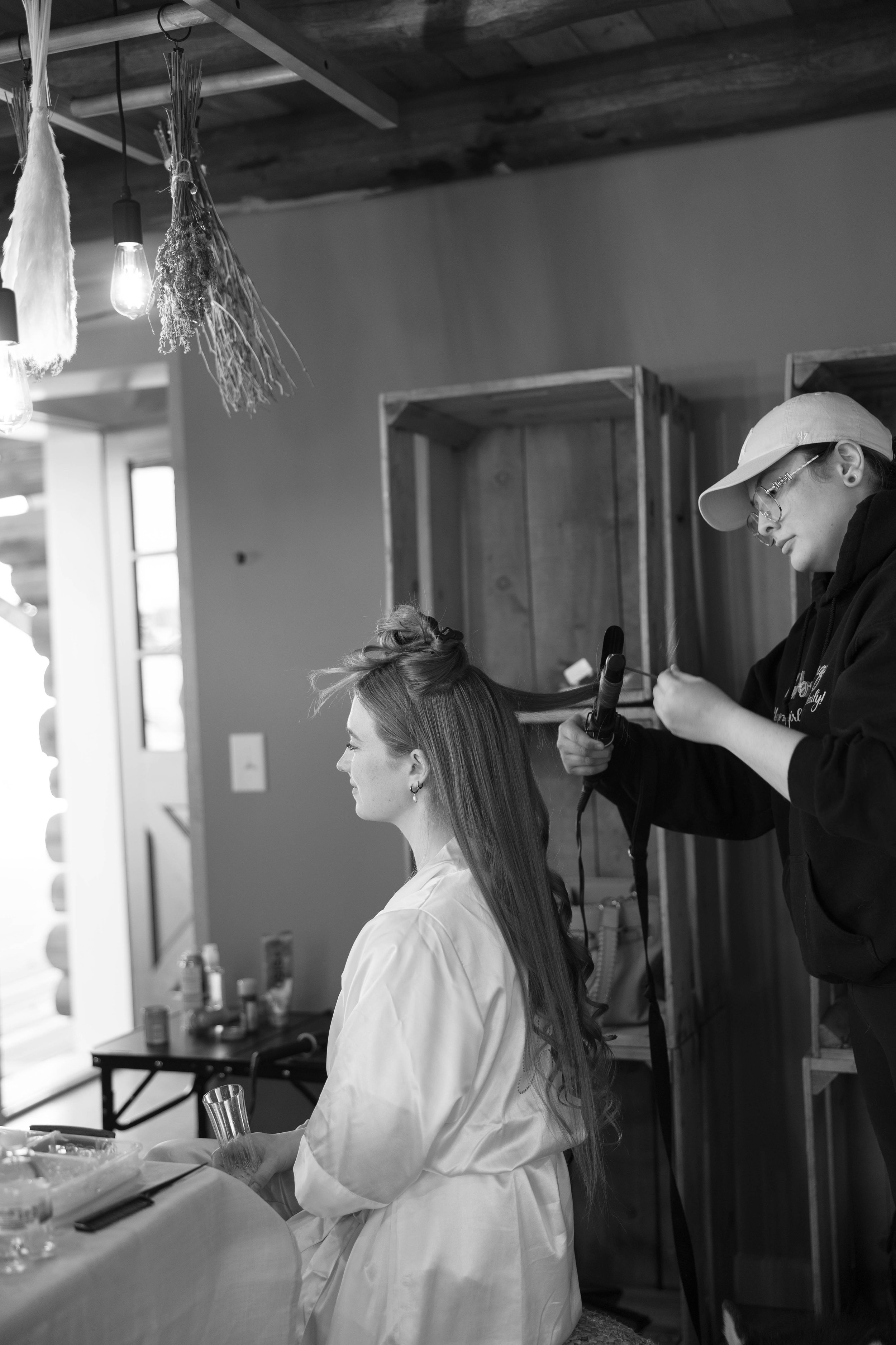 A woman getting her hair styled by a hairstylist in a rustic room with hanging light fixtures and dried herbs.
