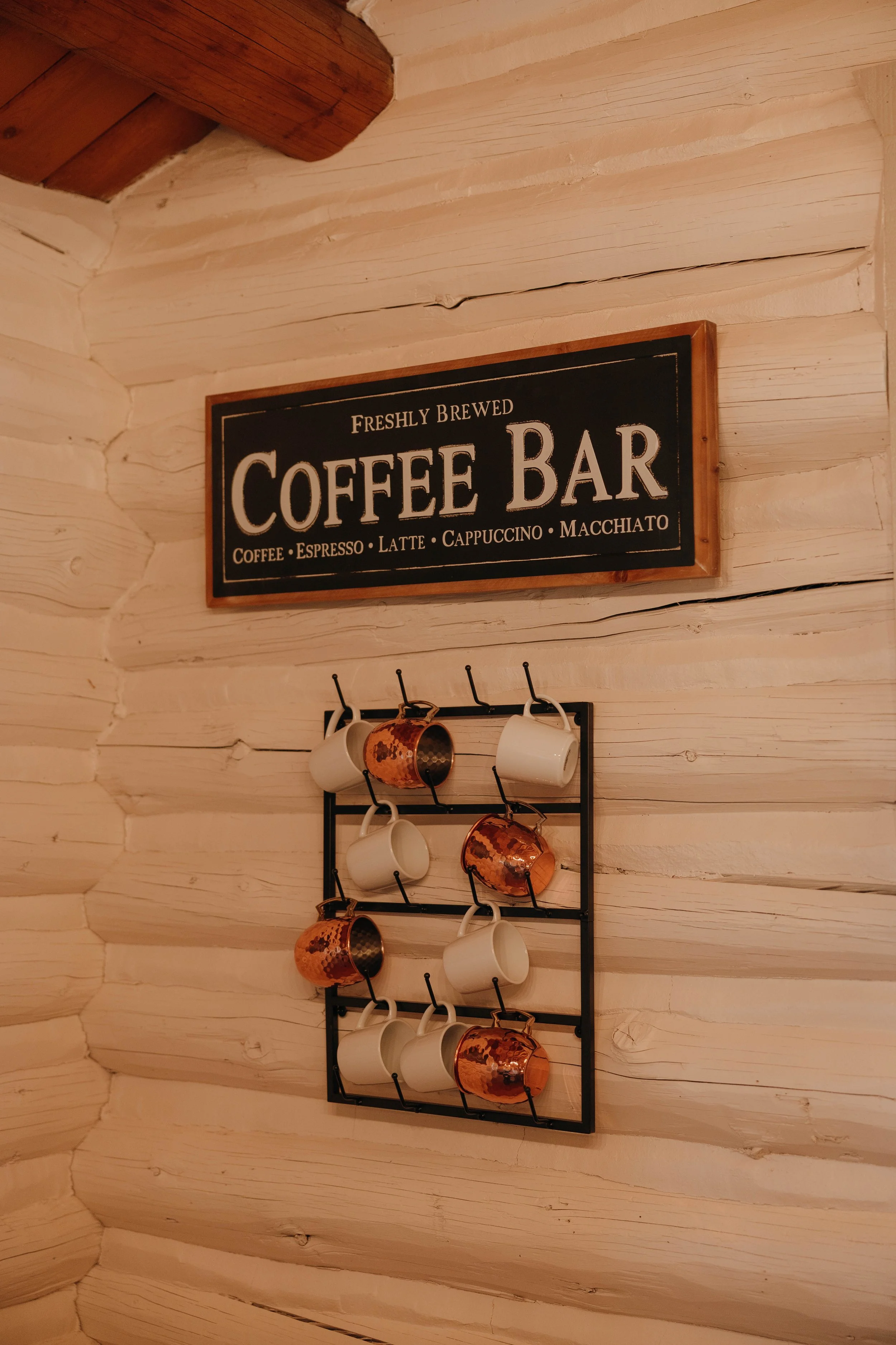 Wooden wall with a sign that reads 'Freshly Brewed Coffee Bar' and a black mug rack holding white and copper mugs.