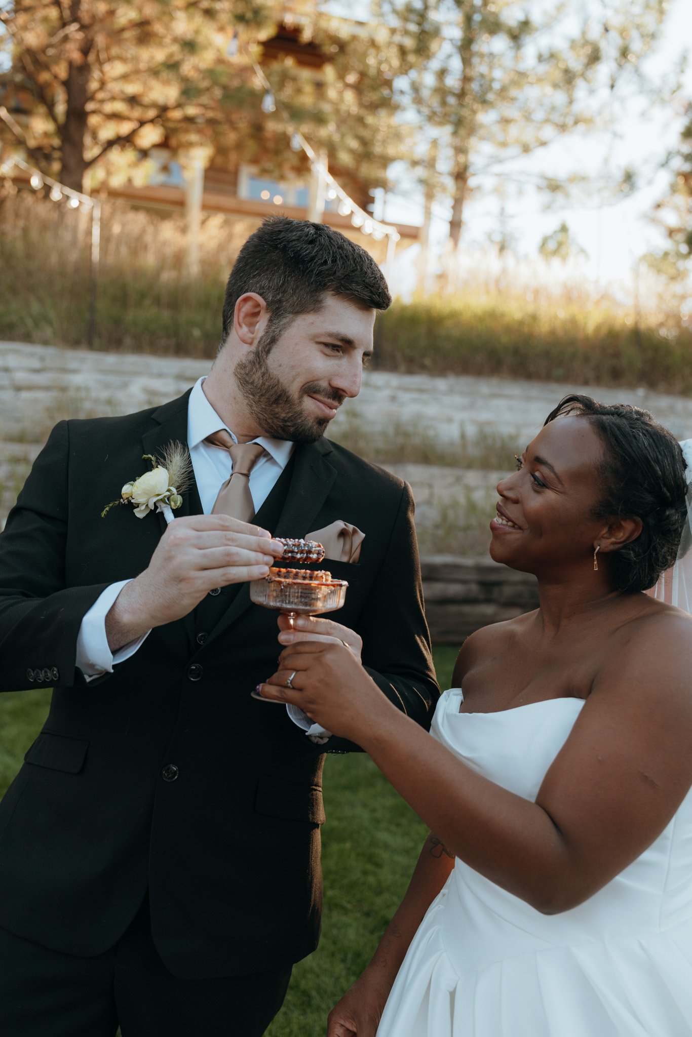A newlywed couple sharing a moment outdoors during their wedding celebration, with the groom holding a decorated cake and the bride smiling at him.