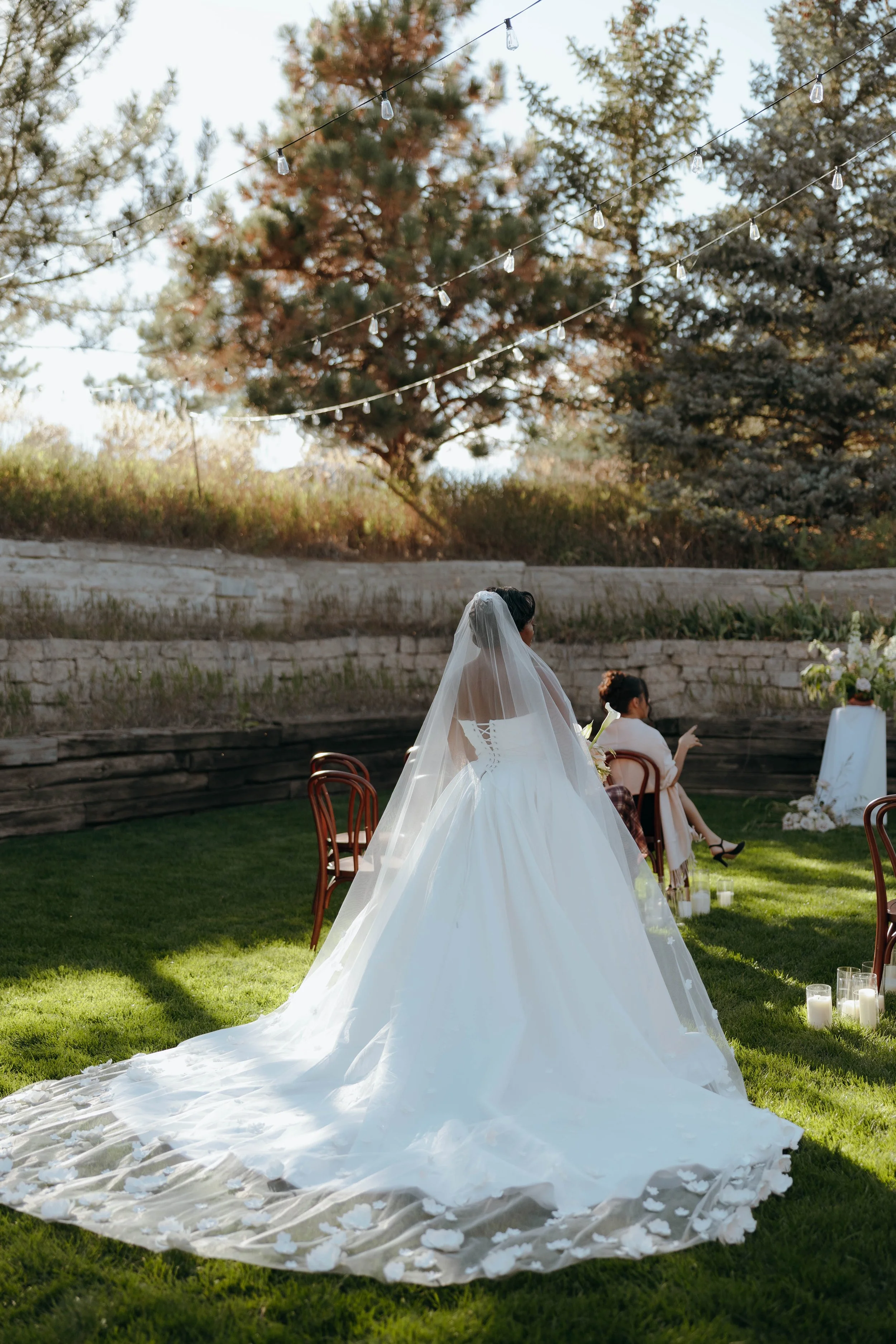 Bride in a white wedding dress and veil standing on green grass at outdoor wedding ceremony with chairs, candles, and floral arrangements, trees, and string lights in the background.