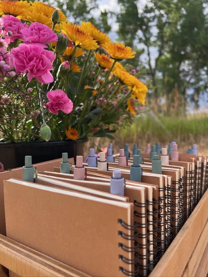A wooden organizer holds spiral-bound notebooks with pastel-colored tabs, placed outdoors next to a pot of pink, yellow, and orange flowers and green trees in the background.