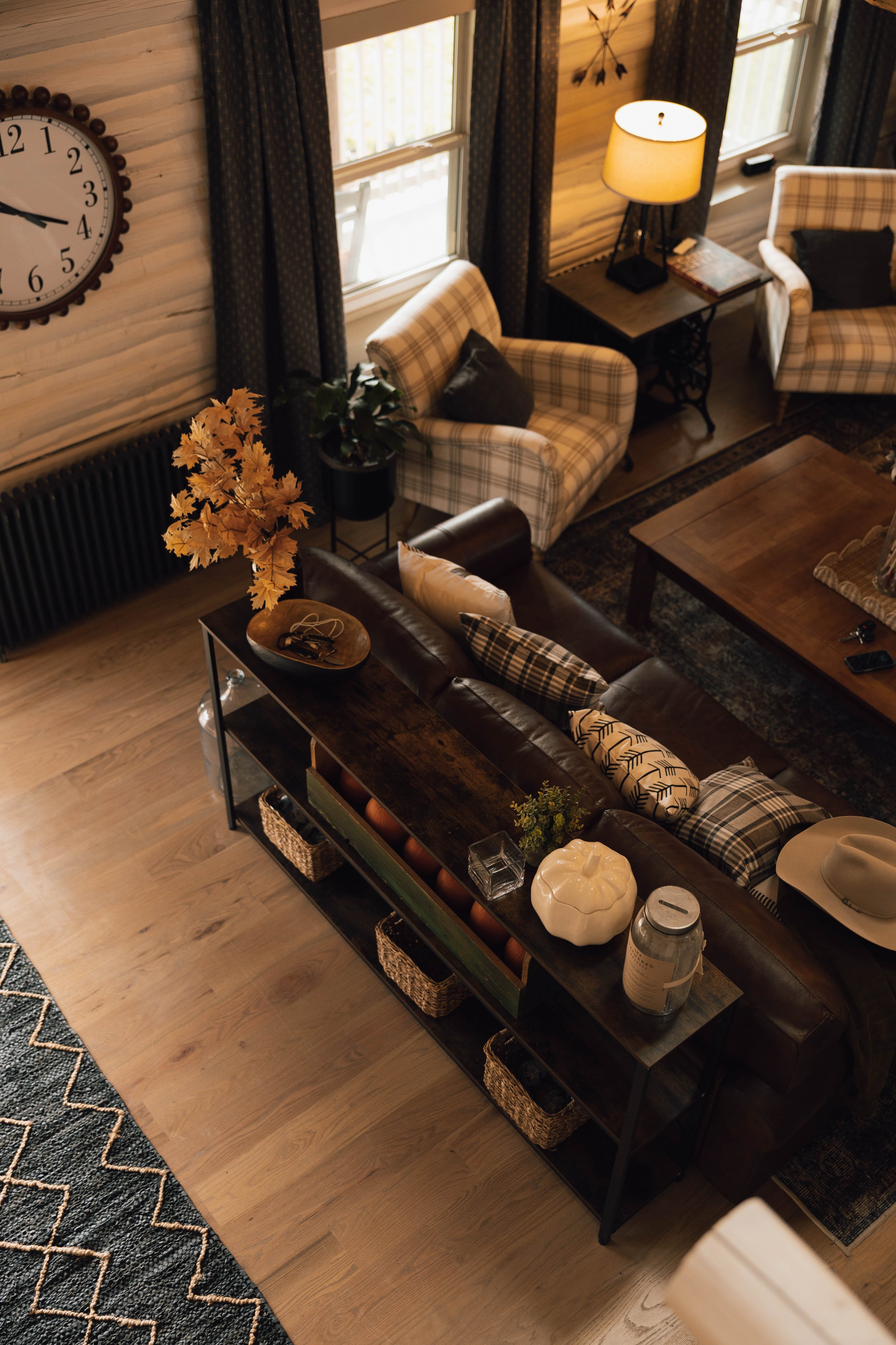 A cozy living room with wooden walls, large windows, plaid armchairs, a leather sofa with pillows, a wooden coffee table, and a side table with a lamp, clock, and decorative items.