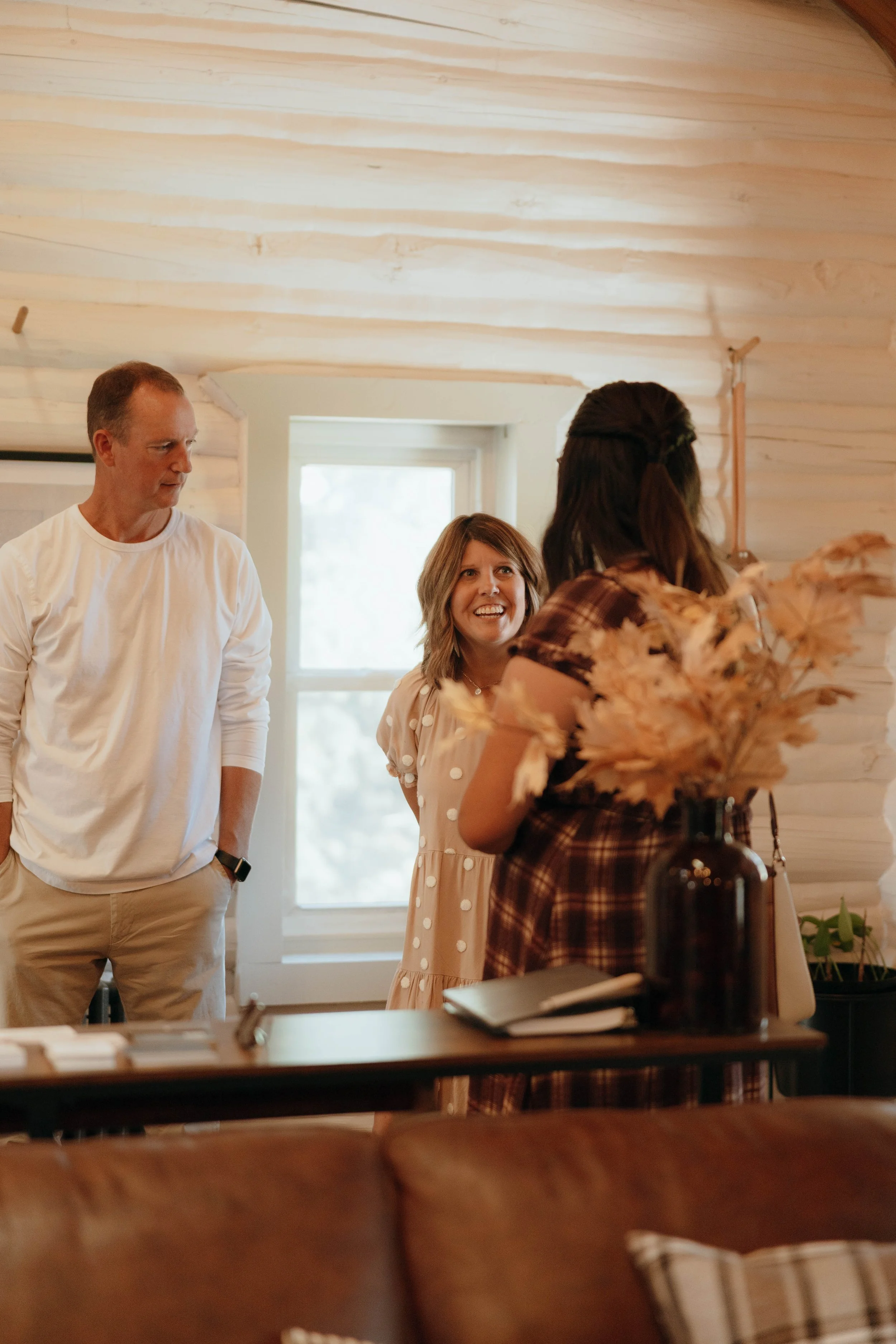 Three people standing and talking inside a cozy, wood-paneled room, with a window in the background and a table with a dark vase filled with dried flowers in the foreground.
