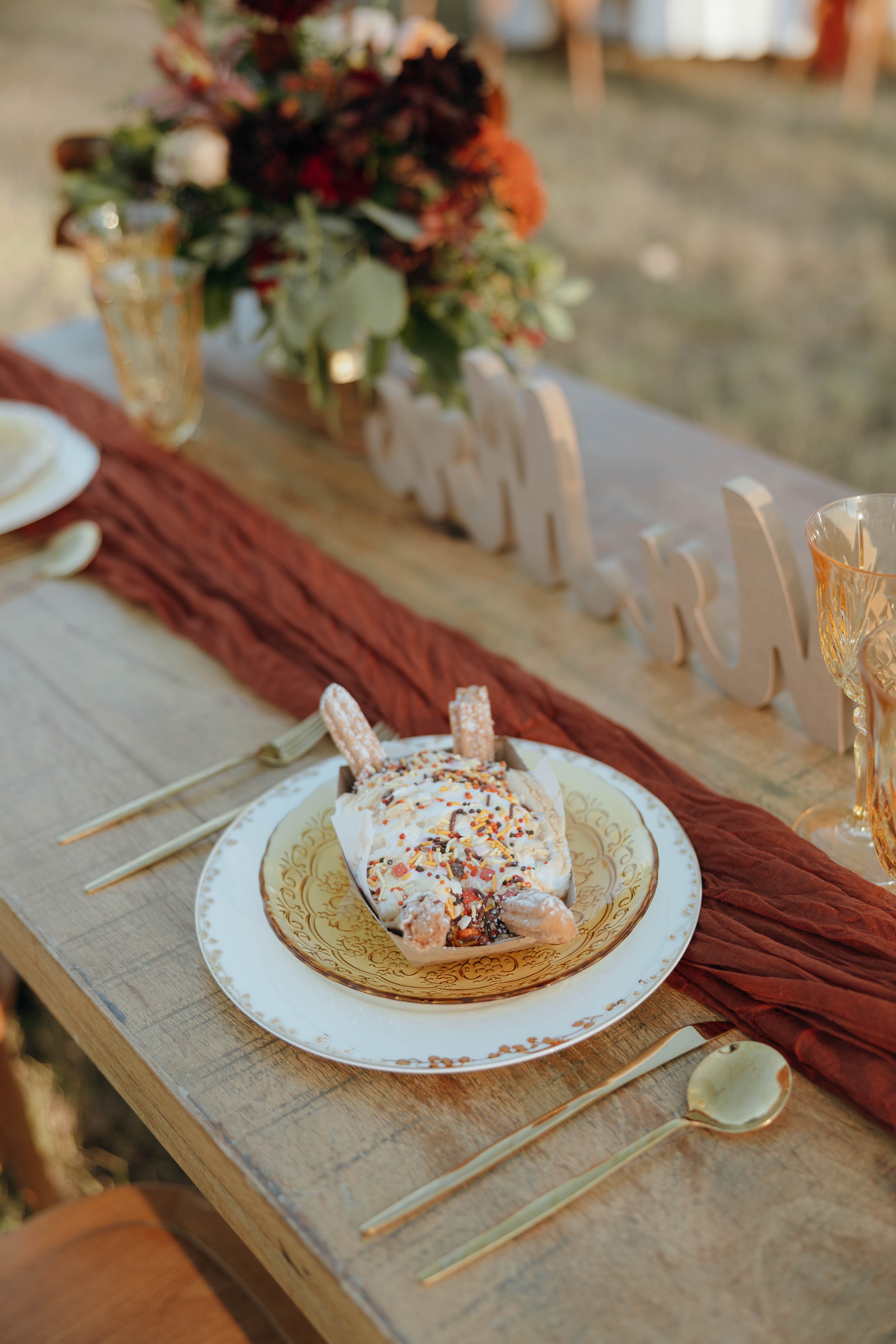 A slice of cake with sprinkles on a decorative plate, set on a rustic wooden table with gold utensils, a brown table runner, and a floral centerpiece in the background.