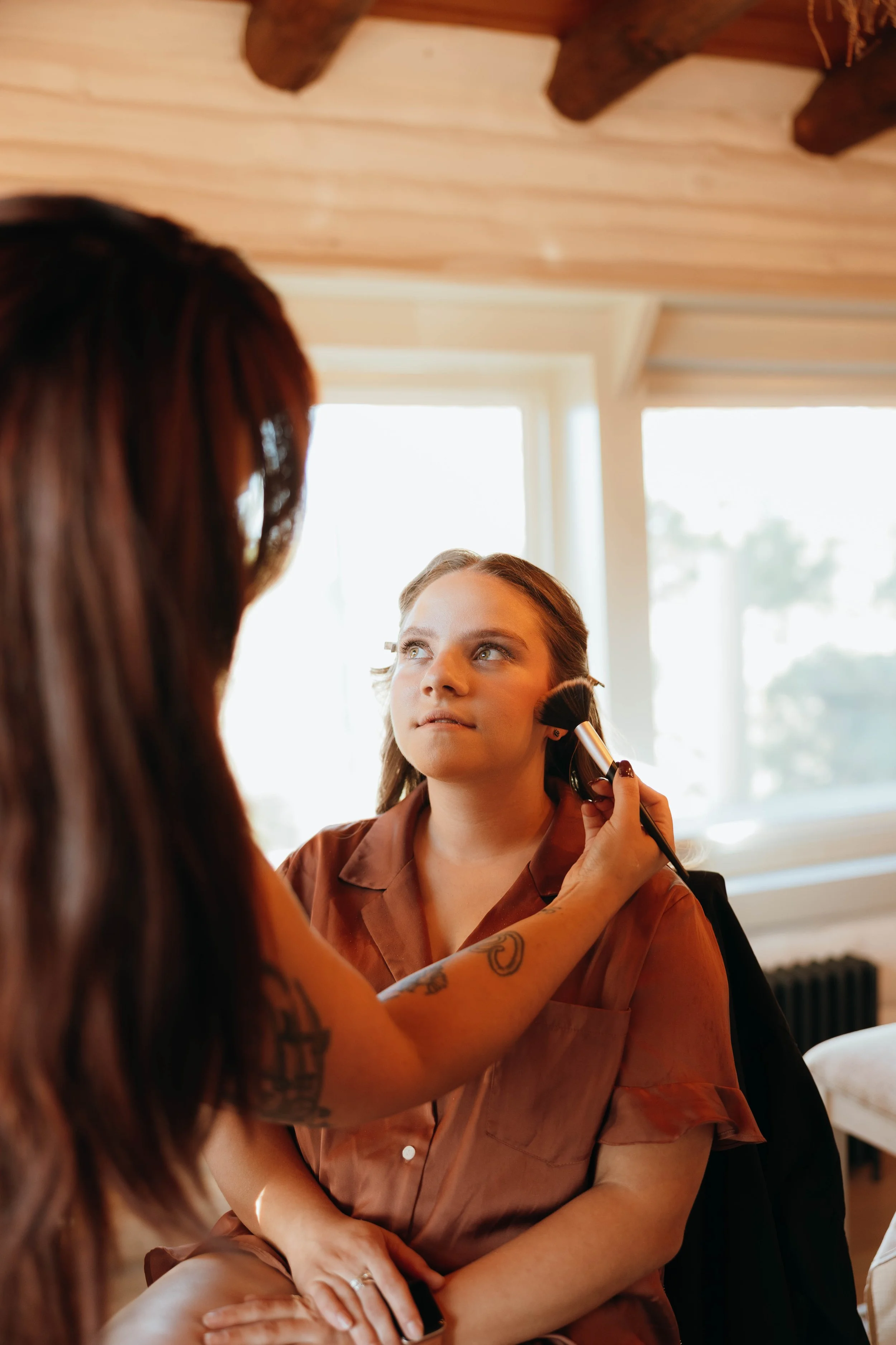 Makeup artist applying makeup to a woman seated indoors near large windows.