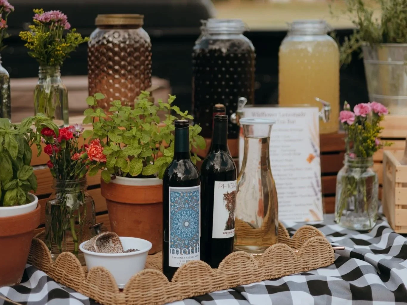 A display of bottled beverages, potted plants with pink and red flowers, jars with honey or syrup, a glass jug filled with a light yellow drink, and a menu on a black-and-white checkered tablecloth.