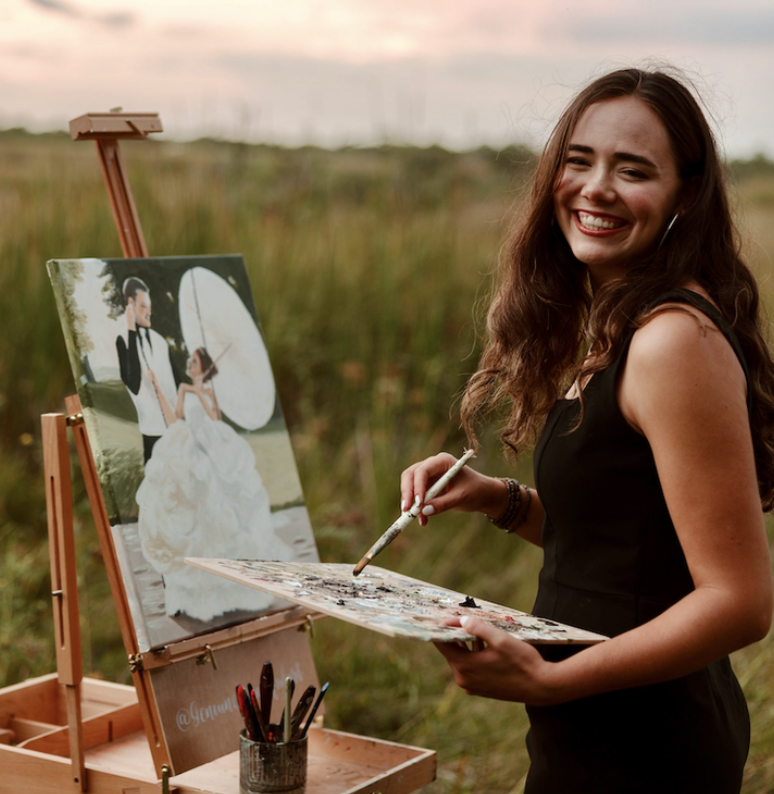 A woman smiling while holding a paintbrush and palette, standing outdoors in a grassy field, painting on a canvas of a couple in wedding attire, with a sky at sunset in the background.