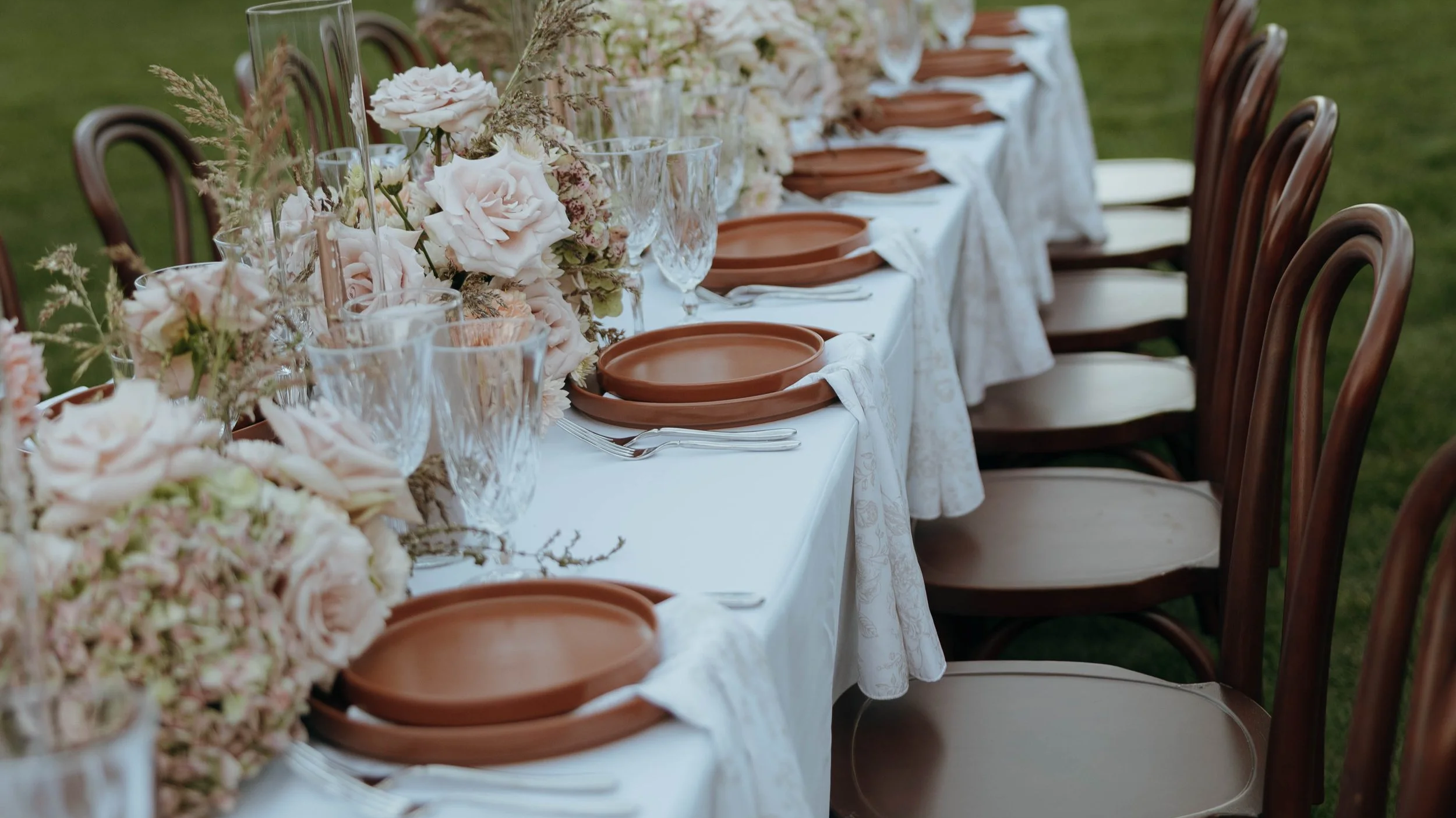 A long banquet table set for an outdoor event, decorated with white tablecloths, pink and cream floral centerpieces, glassware, and brown plates. Wooden chairs are arranged along the sides of the table.
