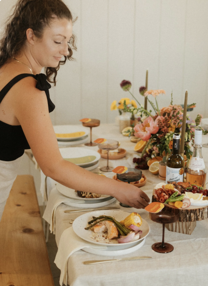 A woman serves herself food at a buffet table decorated with flowers, fruits, and drinks.