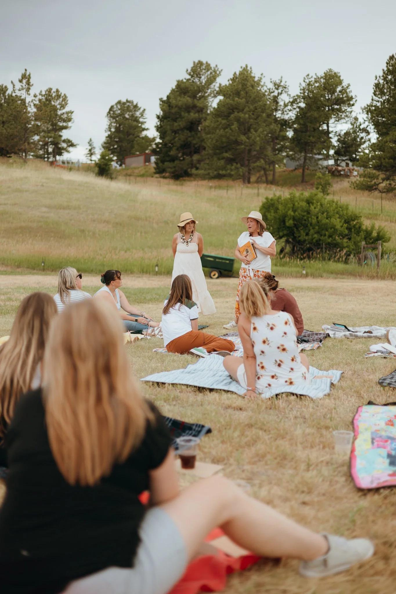 People sitting on blankets outdoors listening to a woman reading, in a grassy field with trees in the background.