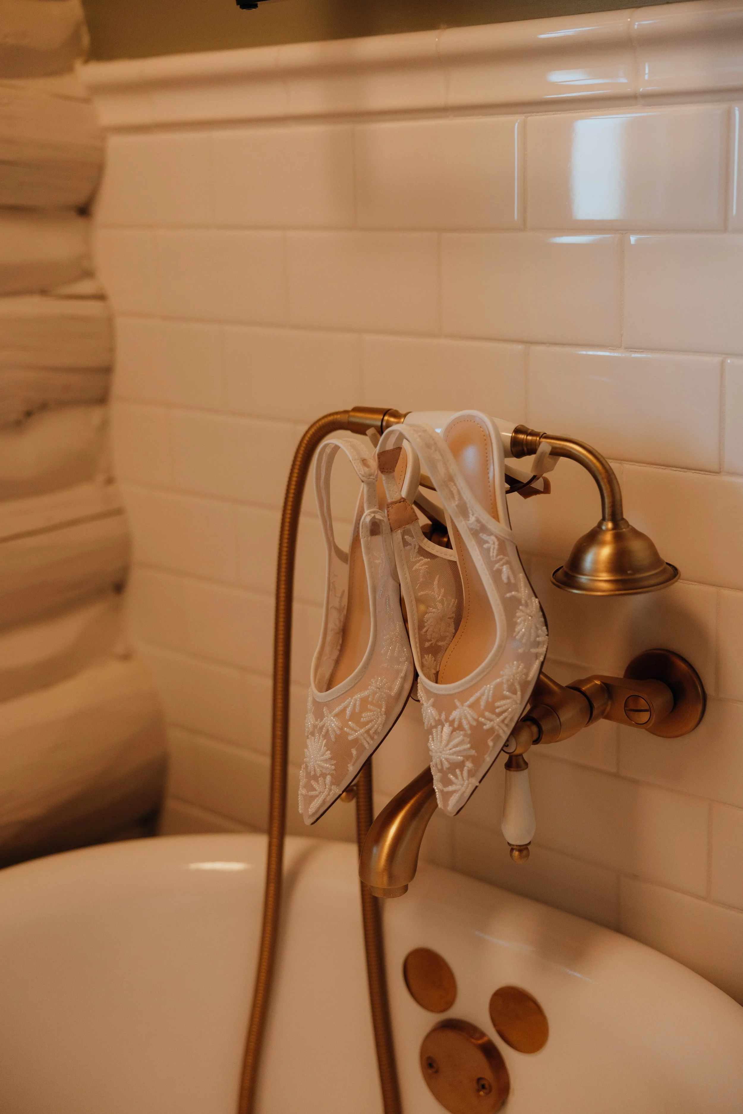 Pair of white embroidered high-heeled shoes hanging from a bathroom faucet with a vintage-style wall-mounted showerhead.