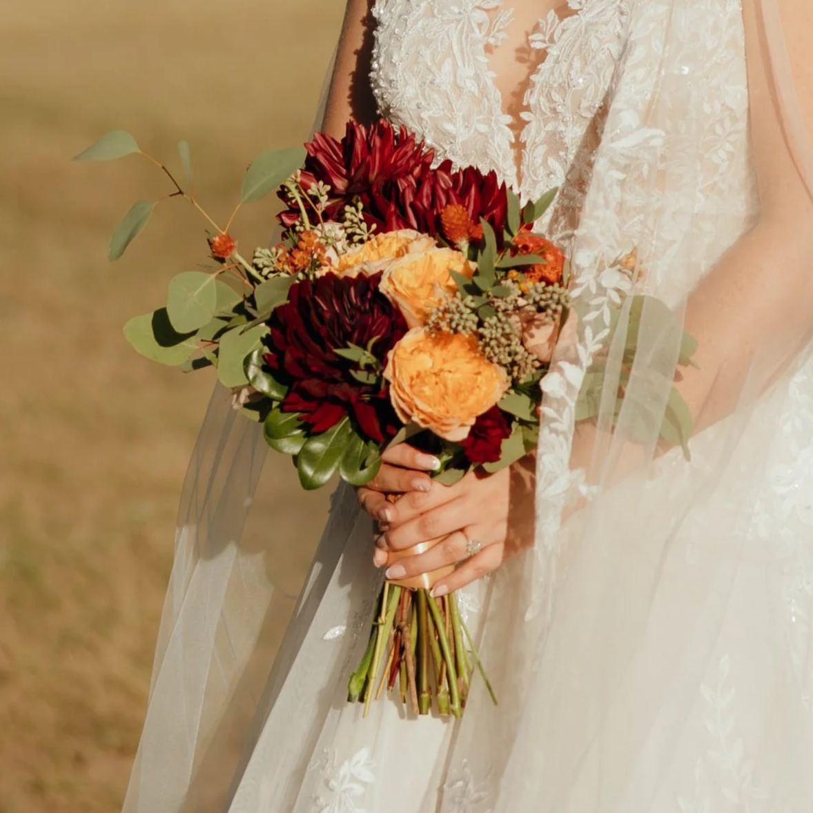 Woman holding a bouquet of red, orange, and peach flowers with green leaves, in a wedding dress.