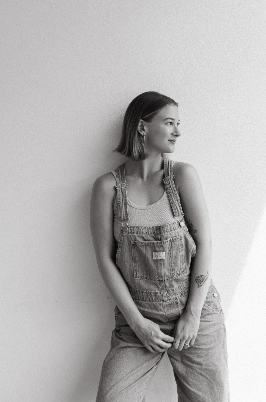 Black and white photo of a woman with shoulder-length hair, wearing overalls over a tank top, standing against a plain wall, looking to her right with a slight smile.