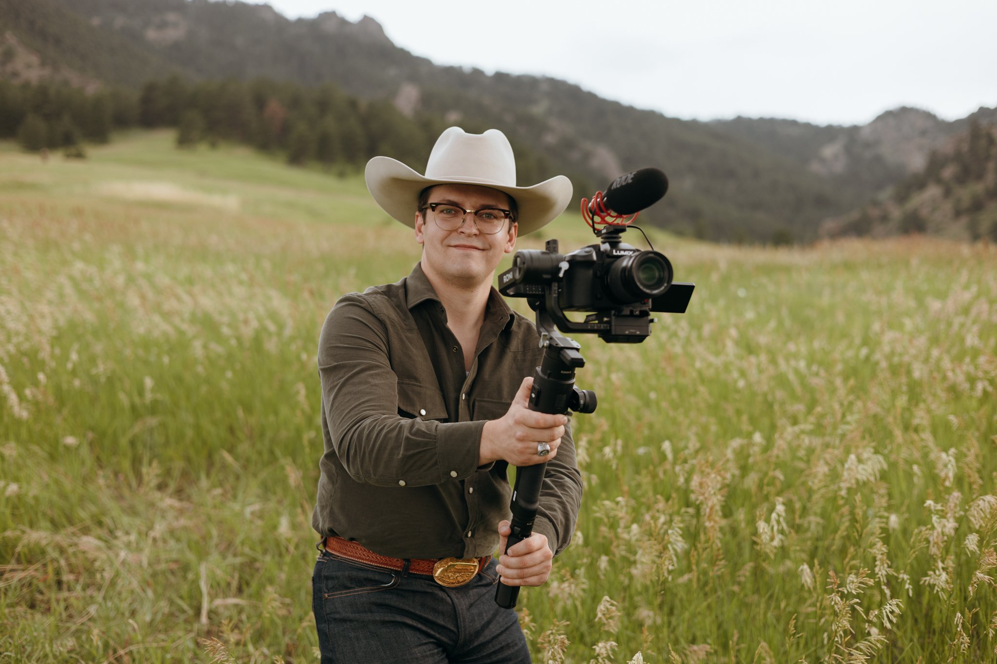 A man wearing a cowboy hat and glasses holding a camera stabilizer with a camera attached, standing in a grassy field with mountains in the background.