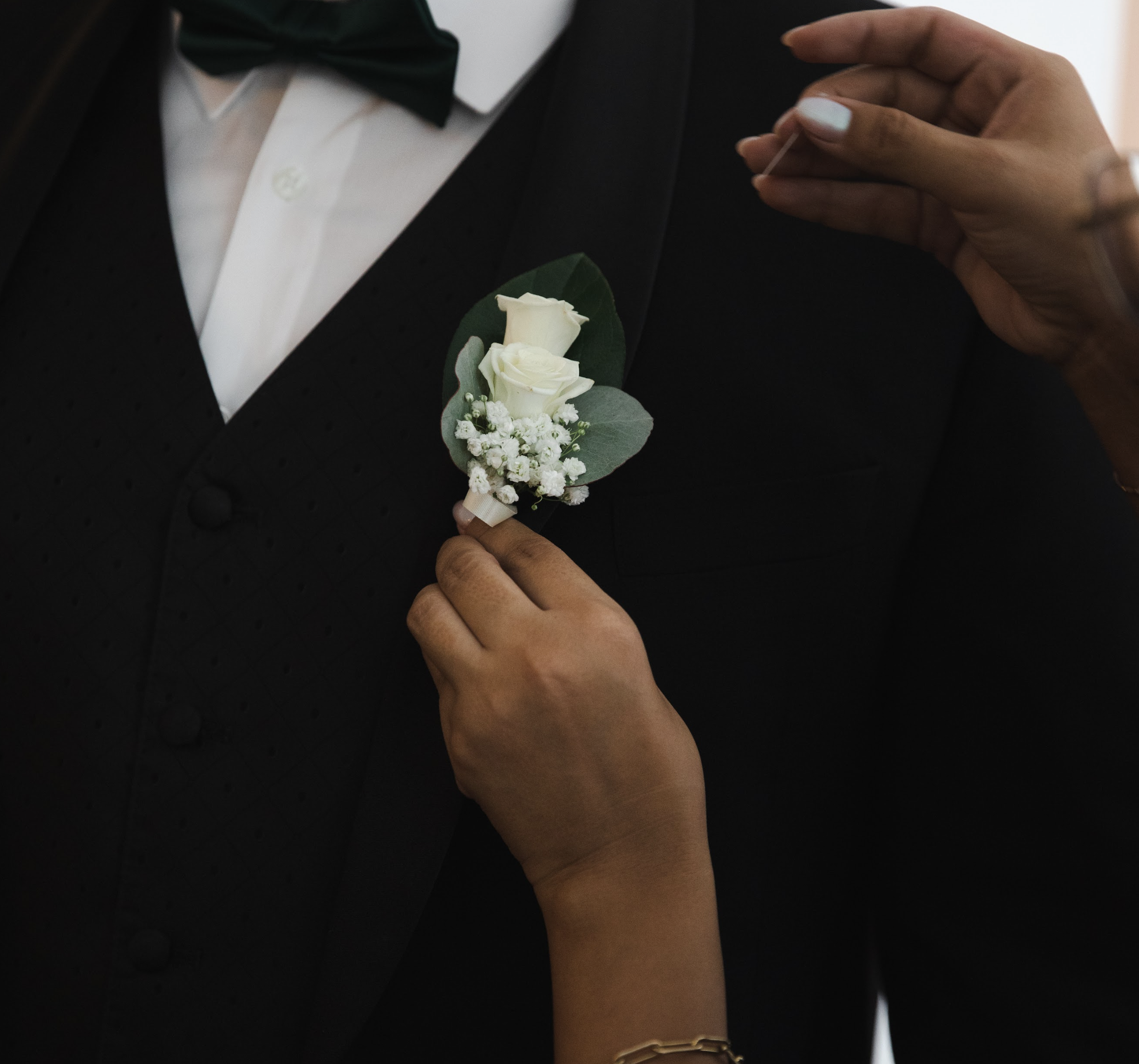 Person pinning a white boutonniere with roses and baby's breath onto a man's black tuxedo.