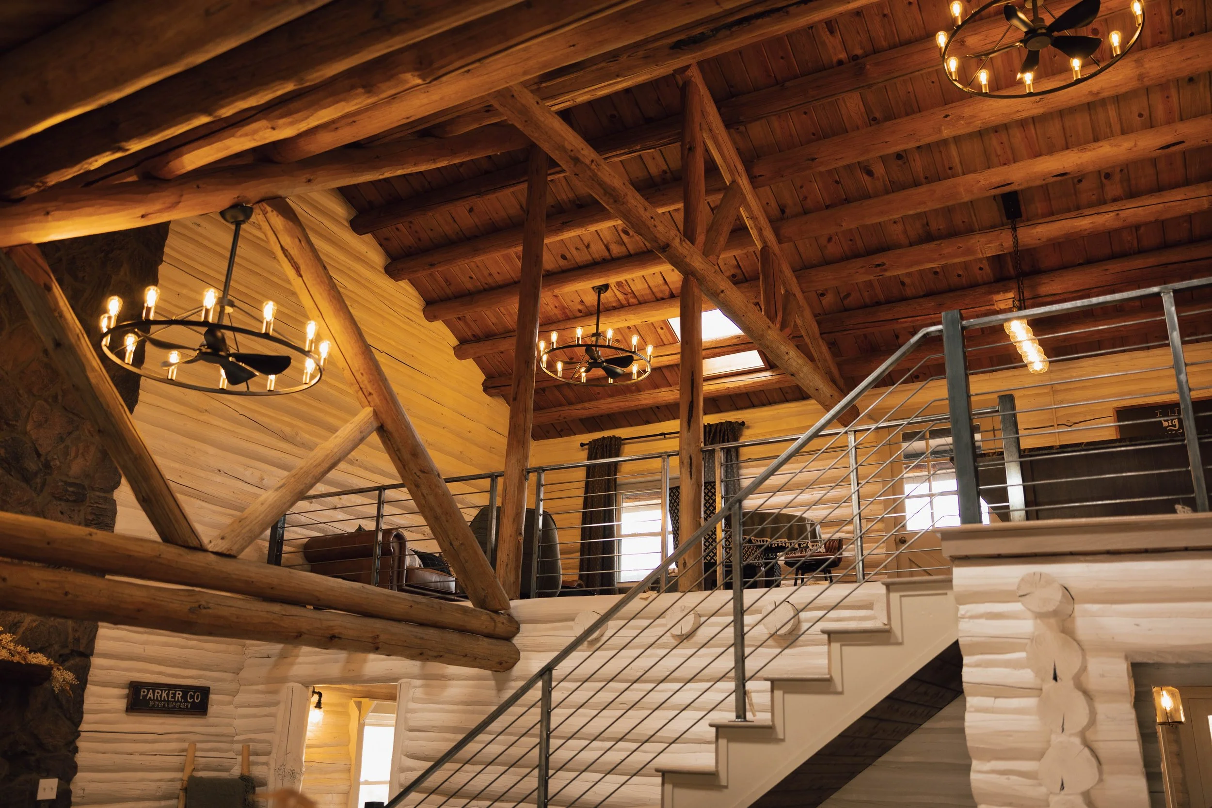 Interior of a rustic cabin with wooden beams, chandelier lighting, and a staircase leading to a balcony with metal railing. The upper level has furniture and windows, and the walls are made of log and stone.