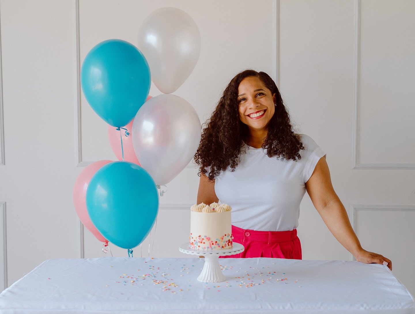 A woman with curly hair smiling in front of a table with a white cake decorated with colorful sprinkles. Behind her are blue, pink, and pearl-colored balloons. The woman is wearing a white shirt and pink pants or skirt, celebrating at a party.