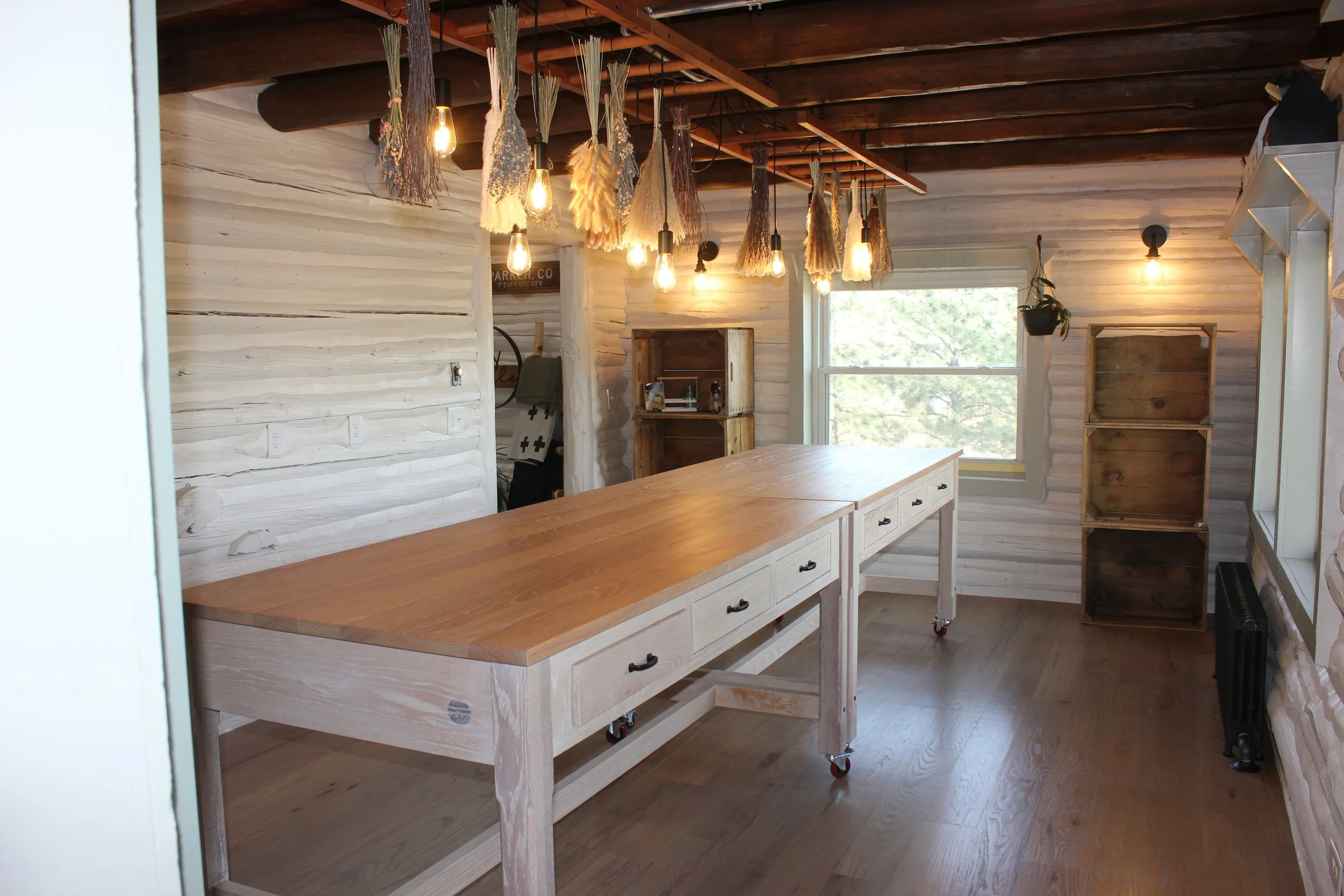 Interior of a rustic room with white wooden paneled walls and a wooden table on casters. Hanging above are multiple bundles of dried herbs or flowers with Edison bulb light fixtures. A large window on the wall lets in natural light, and there are open wooden shelves and a small potted plant near the window.