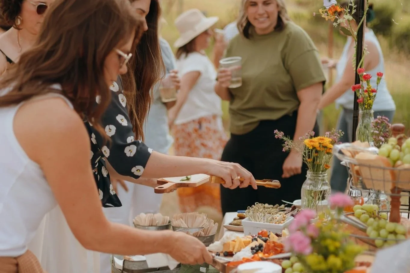 Group of women at an outdoor gathering, serving food from a table decorated with flowers, grapes, and various appetizers.