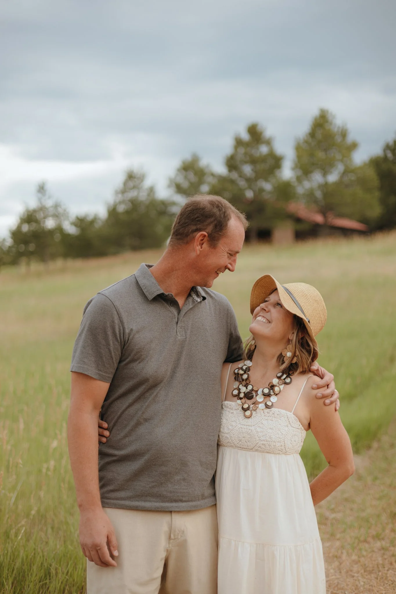 A happy couple standing in a grassy field, smiling and looking at each other, with trees and a cloudy sky in the background.