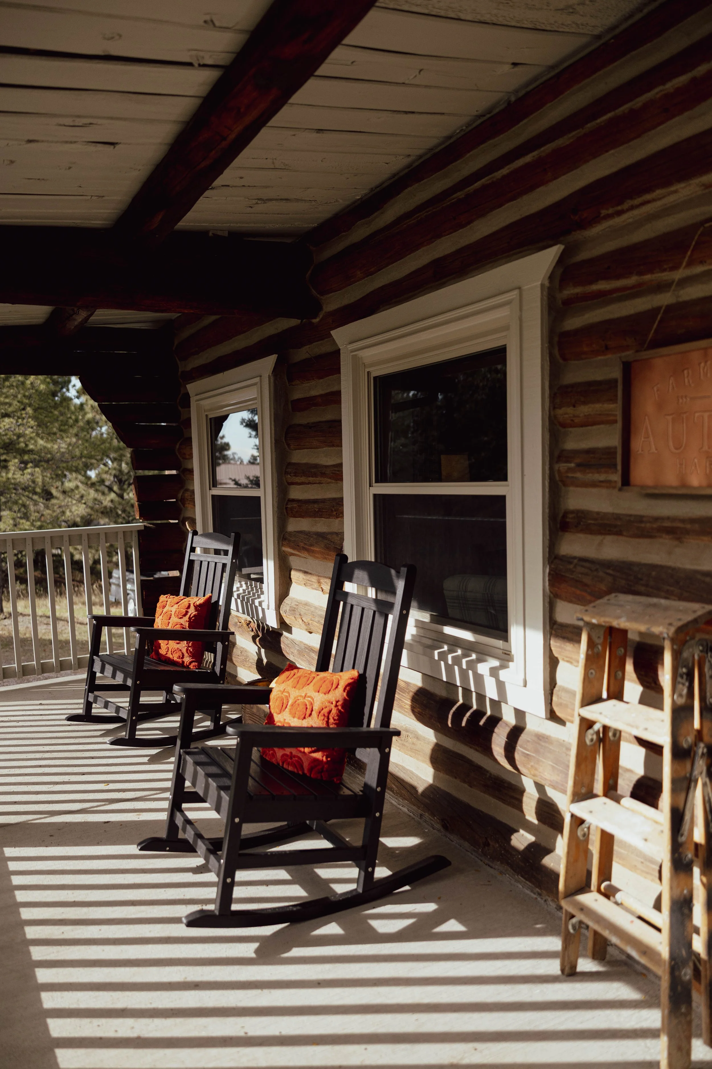 Two black rocking chairs with orange pillows on a porch with diagonal striped shadows. The porch has a wooden railing and log cabin walls with two windows. There is a wooden ladder on the side.