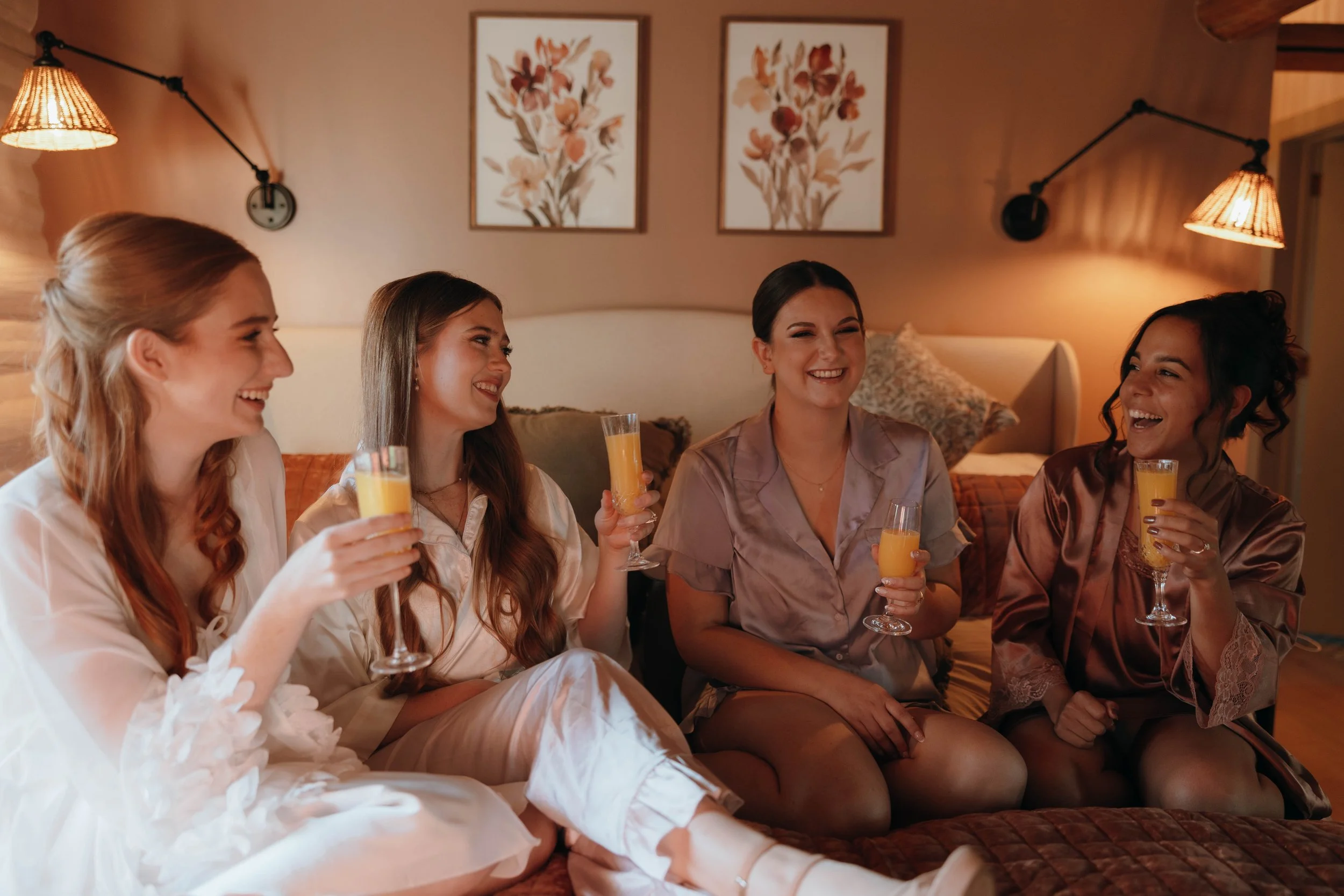 Four women in pajamas sitting on a bed, holding glasses of orange juice, and smiling at each other in a cozy, warmly lit bedroom.