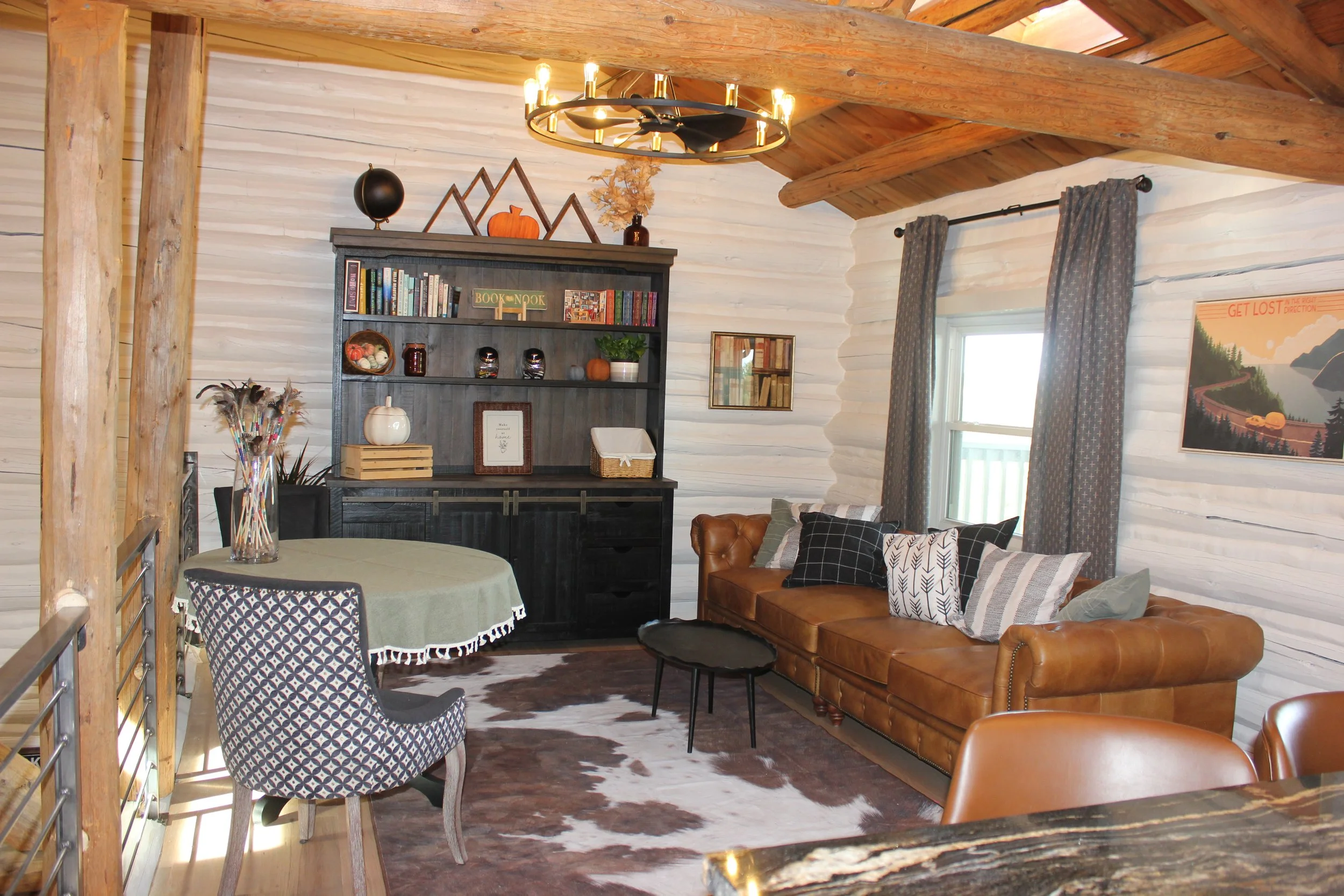 Living room with white wood-paneled walls, a brown leather sofa with assorted patterned pillows, a black bookshelf decorated with pumpkins and books, a round table with a green tablecloth, patterned and solid armchairs, a cowhide rug, a window with patterned curtains, and a wooden beam ceiling with a modern chandelier.