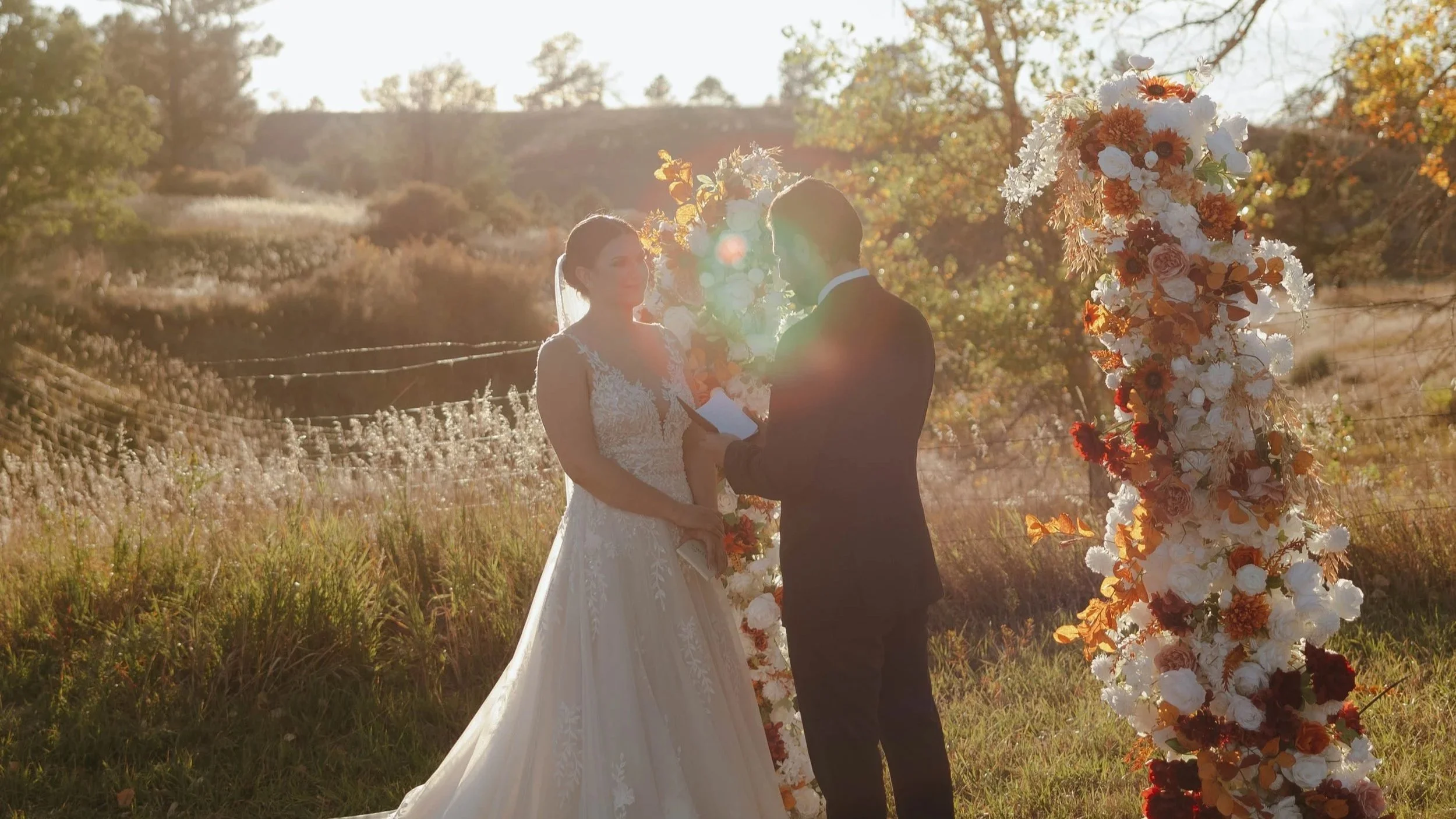 A bride and groom stand outdoors at sunset during their wedding ceremony, exchanging vows in front of a floral arch with orange, white, and red flowers.