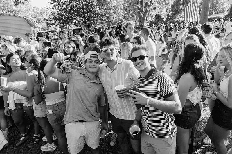 A large outdoor gathering of young adults at a festival or fair with trees and flags, some people holding drinks, smiling and enjoying the event.