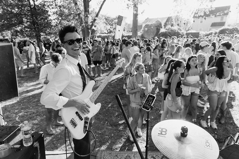 Young male musician with sunglasses playing electric bass guitar at outdoor event with a large crowd of young women and people in the background.