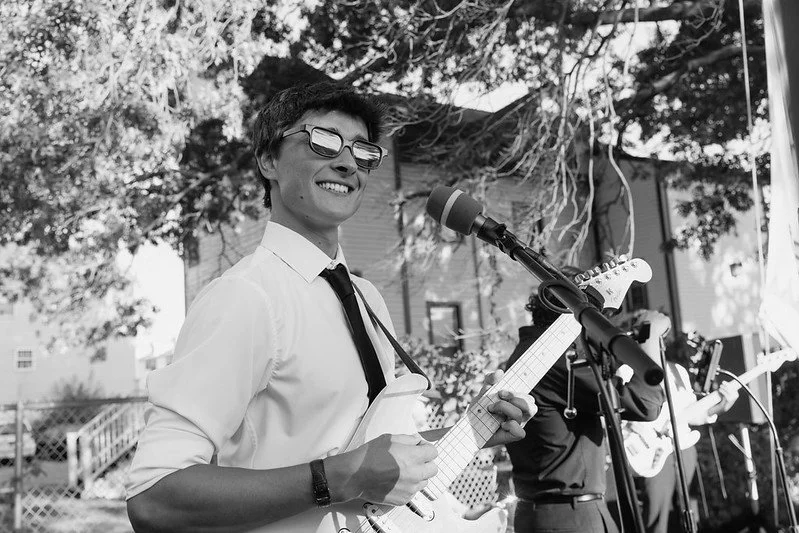 A young man smiling while playing an electric guitar at an outdoor event, wearing sunglasses, a white shirt, and a black tie.