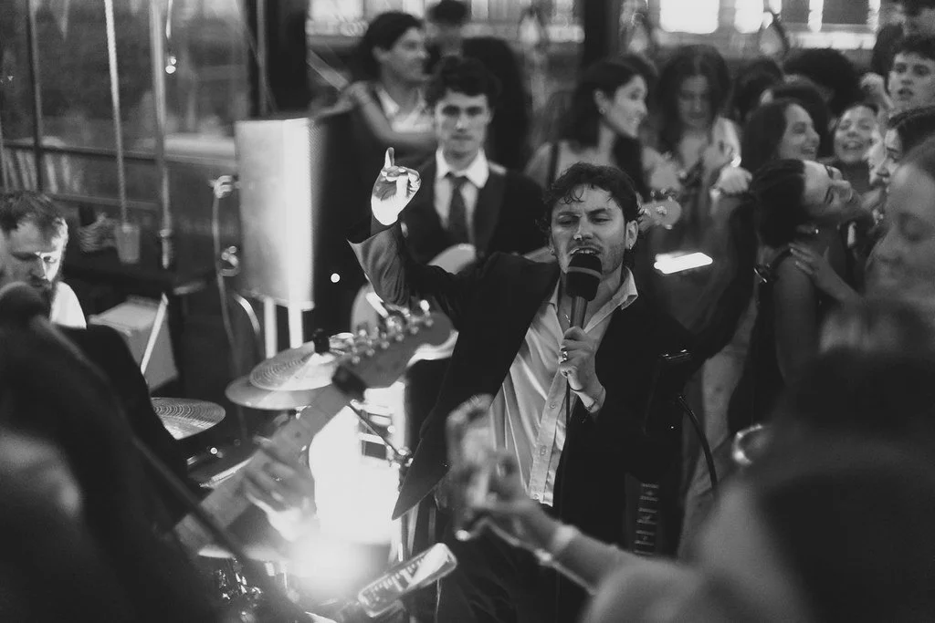 A man singing into a microphone at a lively party with a group of people in the background.