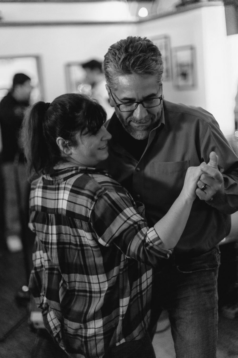 A man and woman are dancing together in a social setting, smiling and holding hands, with other people in the background.