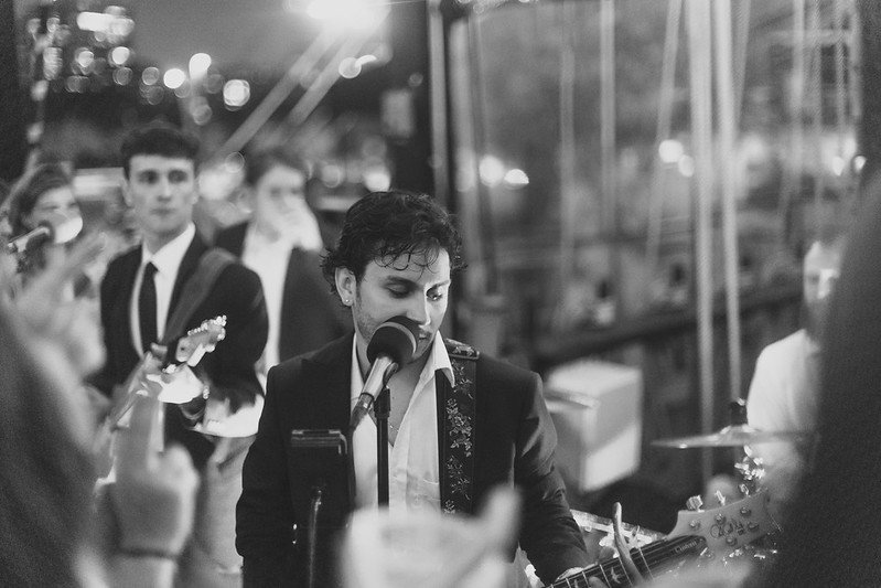 A man with dark, curly hair singing into a microphone while playing an electric guitar at a social gathering or party. Several people, some wearing masks and formal attire, are observing him, with a cityscape visible through the windows in the backgr