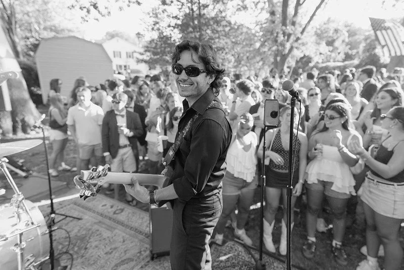 A male guitarist with curly hair and sunglasses performing at an outdoor concert, with a crowd of diverse women and men watching and enjoying the music.