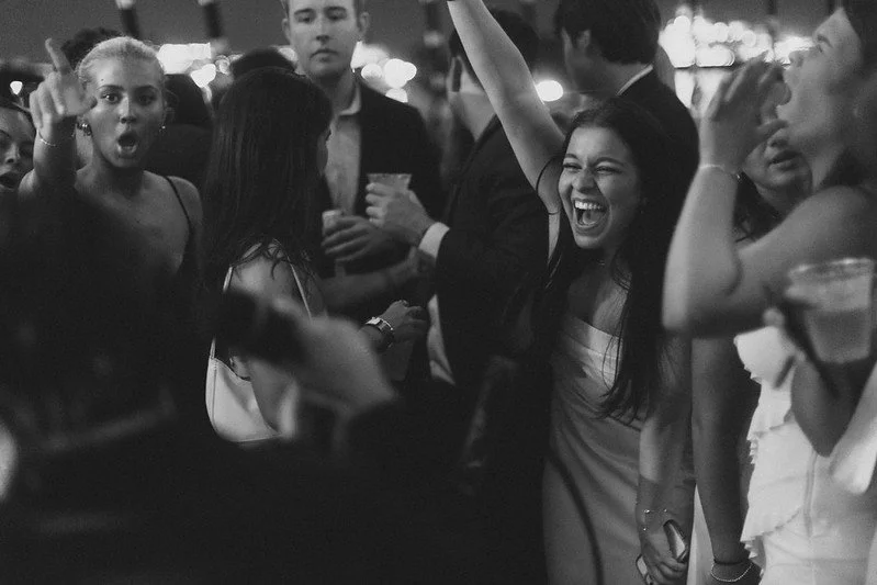Group of young adults at a lively party, some are dancing and laughing, holding drinks, in a dimly lit setting.
